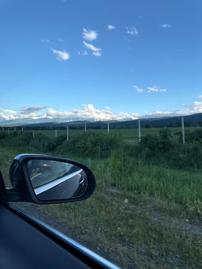 The image, taken from inside a vehicle, shows a view through the side window and the side-view mirror. The side-view mirror reflects the sky, clouds, and the side of the car. Outside the car, a landscape of green fields and vegetation is visible. In the background, a line of trees and mountains can be seen under a bright blue sky with scattered white clouds. A fence line with posts runs horizontally across the middle ground. The overall scene depicts a rural or semi-rural environment. There are no people or specific landmarks to identify the location within the city of Alagir, Russia.