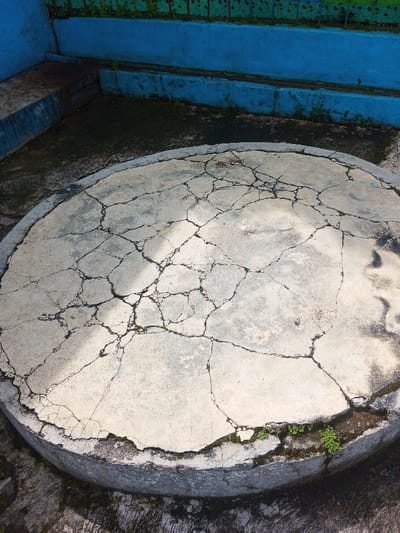 The scene, located in Cipendawa, Indonesia, depicts an outdoor concrete environment under ambient daylight. A prominent, circular concrete slab, approximately 2.5 meters in diameter, occupies the central and foreground area. The slab's light gray surface is extensively fractured with numerous dark, irregular cracks, some containing small green vegetative growths (moss/weeds). The slab's perimeter is slightly elevated, resting on a darker, textured concrete ground. In the background, a multi-tiered concrete structure painted bright blue is visible. This structure includes a lower horizontal base, a vertical wall, and an upper horizontal ledge. The blue paint shows signs of weathering and dirt. Green foliage is present on the uppermost ledge. No individuals or active subjects are depicted.