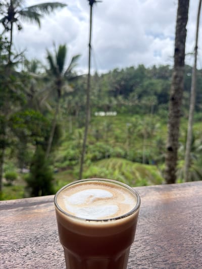 An outdoor shot shows a glass of coffee on a wooden surface with a blurred background of rice terraces and foliage, likely in Tegal Suci, Indonesia. The coffee has latte art, a heart-like shape. The wooden surface has visible grain. The background consists of a verdant landscape featuring rice terraces, trees, and foliage. The sky appears overcast.