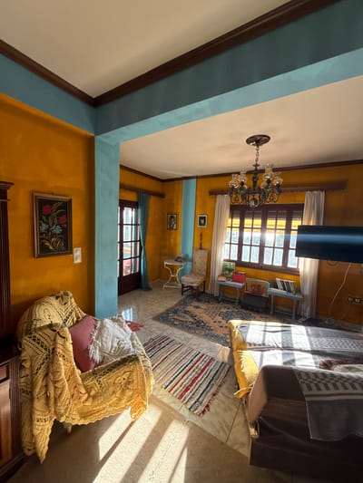 This image depicts an interior residential room, likely a living area, located in Aegina, Greece. The room features walls predominantly painted bright yellow, contrasting with teal-colored structural beams and accent walls. The ceiling is white with dark brown trim where it meets the walls and beams. The floor is tiled with light-colored square tiles, partially covered by multiple rugs, including a large patterned rug with blue and beige tones, and a smaller striped rug.

In the left foreground, an armchair is covered with an intricately crocheted yellowish blanket, holding a pink pillow and a white textured pillow. To the left of the armchair, a dark wooden cabinet or wardrobe is partially visible. A framed artwork featuring red and blue floral motifs is mounted on the yellow wall above a double white light switch.

In the mid-ground, a dark brown wooden door with multiple glass panes is visible, slightly ajar or leading to another space, flanked by a teal curtain. To its right, a small white metal round table is positioned next to a patterned chair, and a framed portrait hangs on the yellow wall above them.

The right side of the room features a large multi-paned dark brown wooden window, through which bright sunlight streams, casting distinct parallel shadows across the floor and furniture. White curtains are drawn to either side of the window. Below the window, two small light blue wooden tables hold books and other items. A dark-colored flat-screen television is mounted on the wall to the right of the window, with electrical outlets visible beneath it. From the ceiling, a chandelier with multiple clear glass shades and green decorative elements is suspended. A sofa or daybed, partially covered with a grey patterned blanket and a brown blanket, occupies the right foreground. No individuals are present in the scene, and no specific actions or interactions are depicted.