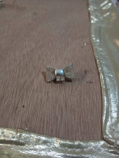 A small, bow-shaped ornament is positioned on a light brown wooden surface in Chandragiri, Nepal. The ornament is metallic, featuring woven golden-toned mesh elements forming the bow's wings, connected by a central, smooth, matte, silver-colored, rounded rectangular band. Below this band, two square settings are visible, each containing a clear, faceted stone. The wooden surface displays vertical graining and minor surface marks. A golden, textured, woven fabric strip borders the scene along the right and bottom, partially enclosing the wooden substrate.