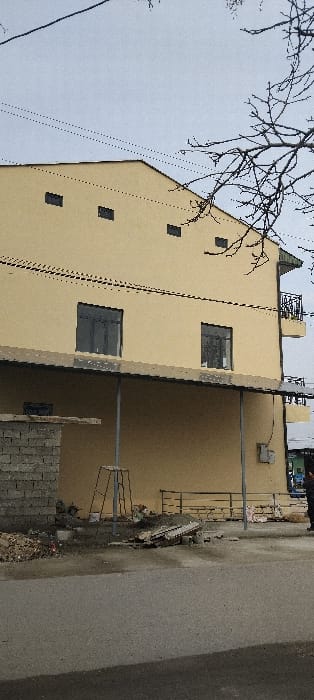 This image depicts a two-story building in Aktash, Uzbekistan, under an overcast sky. The building's facade is light beige. The upper story features a sloped roofline on the left and several small, dark, rectangular openings near the top. Two prominent dark-framed windows are present on the upper story, along with a small, dark-railed balcony on the far right. The lower story is partially covered by a corrugated metal canopy supported by two grey metal poles. Under the canopy, a partially visible window and a grey electrical box are affixed to the wall.
The foreground consists of an asphalt road surface. Adjacent to the building, on the left, is a partially constructed cinder block wall with accumulated dirt and debris at its base. A metal A-frame structure and scattered construction materials, including what appears to be a broken wooden pallet, are visible on the ground. A low, dark metal railing extends along the ground level to the right. A single individual, dressed in dark clothing, is partially visible on the far right, standing on the sidewalk near the building. Overhead, two dark utility lines span the scene against the pale grey sky, with bare tree branches encroaching from the right.