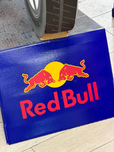 An overhead, close-up, medium shot captures a blue Red Bull branded box. The box is placed on a light-colored wooden floor. On the box, there is the Red Bull logo, which consists of two red bulls charging towards a yellow sun. Below the logo, the words "Red Bull" are printed in large red font. The box appears to be leaning against a large black and grey object which has the texture of a tire. The backdrop consists of a grey, speckled surface. The scene is indoors, with no indication of a specific city or country.