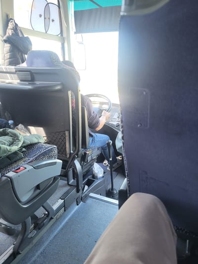 A person wearing jeans and sneakers is driving a bus. The interior of the bus is visible, including the steering wheel, dashboard, seats, and windows. Outside the window, it is bright. Given that this is a bus, the location is likely Orizovo, Bulgaria.