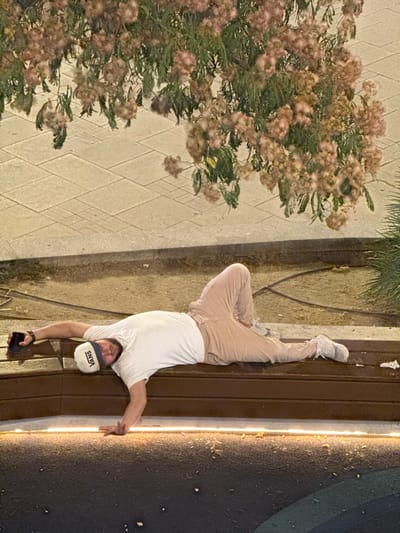 A person is performing a handstand on a wooden bench in a park. The person is wearing a white t-shirt, beige pants, and white sneakers. They are also wearing a white baseball cap with the word "VANS" visible. The person is holding a dark-colored smartphone in their right hand. The background features a tree with pinkish-brown flowers and green leaves, and a paved walkway. The lighting suggests it is nighttime or dusk, with artificial lights illuminating the scene. The ground appears to be a mixture of dirt and dark asphalt with some debris. The image is oriented such that the person appears to be upside down.