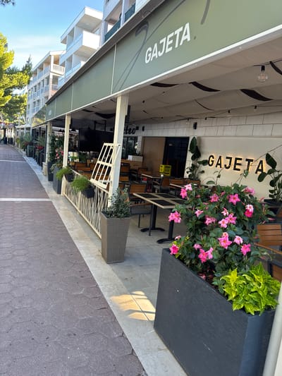 An outdoor seating area for a restaurant called GAJETA is depicted in Makarska, Croatia. The restaurant's name is prominently displayed on its green awning and as illuminated signage on the building's facade. Tables and chairs are set up on a tiled patio, with planters containing vibrant pink flowers and green foliage decorating the space. A walkway paved with hexagonal stones runs alongside the seating area. In the background, a multi-story building with balconies overlooks the scene. The sky is clear and blue, indicating good weather. There are no people visible in the image.