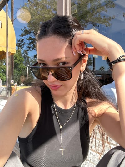 A female individual is positioned for a self-portrait (selfie) in an outdoor commercial setting during daylight hours in Glendale, United States. She has dark, wavy hair, partially pulled back. She is wearing dark brown, flat-top sunglasses with exposed silver rivets; a "Louis Vuitton" monogram is visible on the inner temple. Her eyes are visible above the sunglasses, and her lips are tinted with a light pink gloss, showing a slight smile. She wears a black ribbed tank top. A long, gold-toned rosary-style necklace with a crucifix pendant hangs around her neck. A small golden star-shaped adhesive is present on her left upper arm/shoulder. Her left hand, adorned with a silver ring on the fourth finger and light pink manicured nails, rests near her left temple, partially supporting the sunglasses. On her right wrist are multiple black beaded bracelets, some with gold-colored accents. Drop earrings with clear, rectangular faceted stones are worn. Gray fabric, consistent with pants, is visible at the bottom of the frame.
The background consists of strong sunlight reflecting off a large glass storefront. Visible reflections in the glass include a bright blue sky, green tree canopy, a distinct spherical white light fixture, a yellow fabric awning, and two faint human figures—one appearing seated, the other less defined. Palm trees are reflected in the upper right portion of the glass. A vertical brown structural beam separates glass panels, and a light-colored brick wall is visible below the reflections. The scene is consistent with an outdoor cafe or restaurant area on a commercial street.