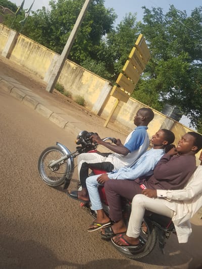 The image shows four young men riding on a single motorcycle on a paved road in Dutsinma A, Nigeria. The rider wears a blue and white shirt, while the three passengers wear a brown robe, a light blue shirt and light coloured pants respectively. None wear helmets or shoes. They appear to be travelling in front of a faded yellow building with a street sign attached to it. The environment is sunny and there are trees visible in the background.