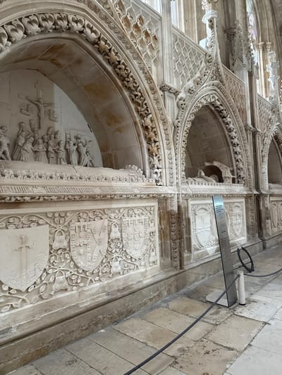 The image displays an interior section of the Monastery of Batalha in Batalha, Portugal, featuring a series of ornate stone tombs integrated into a wall. On the left, a large arcosolium contains a detailed bas-relief carving depicting the Crucifixion of Christ, with Christ on the cross and approximately twelve figures, including mourners and a soldier, gathered below. The tomb's lower facade beneath this relief features a frieze with an inscription, followed by three prominently carved heraldic shields. The leftmost shield depicts a sword, while the two central shields exhibit complex coats of arms surrounded by intricate foliate patterns. To the right, several additional arcosolia are visible, each with highly decorated Gothic arches and recesses containing smaller, recumbent effigies. The surrounding wall is adorned with extensive Gothic architectural details, including tracery, finials, and carved ornamentation. The floor is composed of large, worn stone slabs. A black rope barrier supported by white posts demarcates a viewing area in the foreground, with a tall metallic informational sign positioned behind it to the right. The scene is illuminated, highlighting the intricate stone carvings.