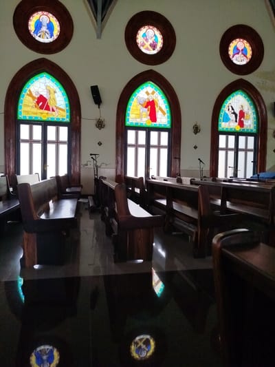 The image shows the interior of a church in K'Rèn, Vietnam, featuring rows of wooden pews and stained-glass windows. The windows depict religious scenes and figures, with arched frames and dark wooden trim. Above the arched windows, smaller circular stained-glass windows are also visible. The walls are painted a light color, and the floor is dark and highly reflective, creating reflections of the windows and pews. Several microphones on stands are positioned between the pews. The overall impression is one of a traditional, well-maintained place of worship.