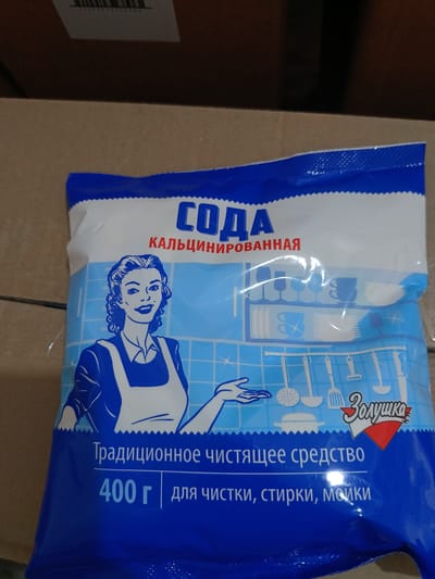 The image shows a close-up of a blue and white bag of "Soda" (sodium carbonate) cleaning powder. The packaging features a retro-style illustration of a woman in an apron, along with kitchen utensils and dishes. Text on the bag indicates it is a "Traditional cleaning agent" and weighs 400 grams, used for "cleaning, washing, scrubbing." A logo with the word "Золушка" (Zolushka, meaning Cinderella) is also visible. The bag is placed on a cardboard surface. The scene does not contain any information that can identify its location within the city of Berdiansk, Ukraine.