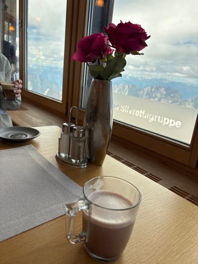The image shows a table setting in what appears to be a restaurant or cafe with a panoramic view of mountains through a large window. In the foreground, a glass mug containing a frothy, chocolate-colored beverage sits on a wooden table. To the left, a grey placemat covers part of the table, and a small, dark grey saucer is visible. A salt and pepper shaker set with a metal rack is positioned to the right of the placemat.

In the middle ground, a metallic vase holds two artificial pink roses. The background is dominated by a large window offering a breathtaking view of snow-capped mountains under a cloudy sky. A white decal with text, partially obscured, reads "Silvrettagruppe," suggesting the location is likely in the Silvretta region, potentially near Ehrwald, Austria, given the mountainous terrain.

A person's hand is visible on the far left, holding a glass of a similar beverage. The overall atmosphere suggests a cozy and scenic dining experience.