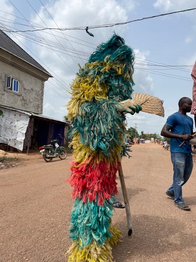 In Agba Umana, Nigeria, a colorful raffia masquerade costume is centered on a brown dirt road. The costume displays horizontal bands of yellow, green, and red raffia. A gloved hand holds a woven fan. A wooden pole acts as a brace, with a person's foot in a sandal visible at the base. To the left, a partially constructed building with a gray exterior, white-framed window, and a dark gray roof is visible. A green motorcycle is parked in front of a building shrouded in white and blue tarp. In the background, a man in a blue shirt and jeans is standing, holding a wooden object. The sky is overcast with visible power lines.
