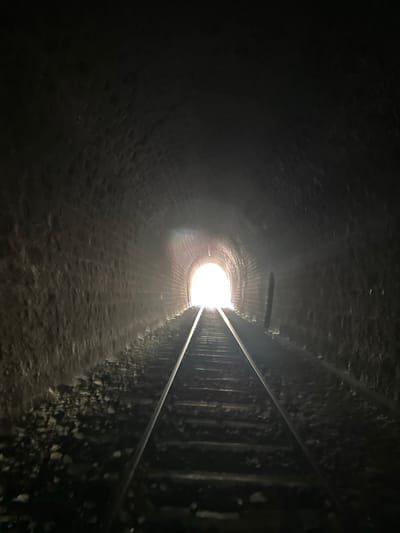 The image captures the interior of a dark brick tunnel with railway tracks running through it. The tunnel walls are constructed from aged, rough-hewn bricks, and the ground beside the tracks is covered in gravel and debris. At the end of the tunnel, a bright, white light emanates, suggesting an exit. The perspective is from a low angle, looking down the length of the tracks towards the light. There are no people or animals visible in the frame. The tunnel is located in Générargues, France, specifically the Pont-du-Gard railway tunnel, which is a disused railway tunnel.