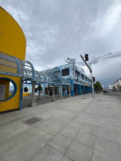 An outdoor, eye-level, wide-angle shot captures a cloudy day in downtown Anchorage, Alaska, USA. The foreground is dominated by a grey concrete sidewalk. To the left, a curved, bright yellow building with a blue trim features a circular cutout and an adjacent covered walkway with a glass roof and blue supports. The walkway leads to a blue building with a stylized white orca decoration on its facade. This building has multiple windows and doors. A sidewalk stretches ahead, leading to a street with a traffic light. Cars are visible on the street, moving away from the camera. The sky is overcast with grey clouds.