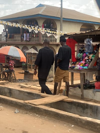 This image depicts an outdoor market scene in Egbedi, Nigeria, characterized by unpaved ground and commercial activity under a cloudy sky. In the foreground, a wide concrete gutter or ditch traverses the scene, with two wooden planks serving as makeshift bridges. Two adult males are walking away from the viewer, traversing the area near the ditch. The man on the left wears a dark tunic over dark trousers. The man on the right wears a black t-shirt, tan shorts, and a black beanie, with a light-colored object visible on his left foot.
To the far left, a man in a patterned shirt and shorts is seated on a wooden stool at a wooden table, partially shaded by an orange and white umbrella. Near him, a dark metal basin rests on the ground. To the far right, a small child in a red top sits on a low concrete ledge. Adjacent to the child, a market stall displays numerous colorful sandals and slippers on a wooden tiered stand, with a red garment hanging behind it.
In the background, a two-story building with a brown corrugated metal roof and blue trim dominates the upper half of the image. The upper floor features a balcony with a white balustrade, from which various clothes are hung. Yellow and black patterned bunting is strung across the balcony. Several arched openings on both floors reveal more merchandise, suggesting shops within the building. Other individuals are visible on the balcony and further within the market.