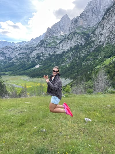 A woman is captured mid-jump in a grassy field with a mountainous backdrop. She is wearing a plaid shirt, denim shorts, sunglasses, and bright pink sneakers. Her arms are extended, with her right hand pointing outwards. The background features steep, rocky mountains covered in green foliage and scattered trees, with a valley stretching out below, dotted with yellow flowers. The sky above is partly cloudy with bright sunlight. The scene appears to be in a rural, mountainous area, likely in the region of Shalë, Albania, known for its natural beauty and dramatic landscapes.
