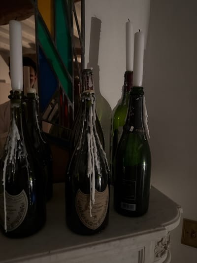 An interior scene captured in Mauléon-d'Armagnac, France, features a collection of approximately seven repurposed glass bottles, primarily dark green champagne bottles (at least two identified as Dom Pérignon Vintage 2009), standing on an ornate white mantelpiece. Several tall, unlit white taper candles are inserted into the necks of these bottles. Significant amounts of solidified white wax drip down the sides of multiple bottles, indicating previous candle combustion. The mantelpiece exhibits carved embellishments on its right side. In the background, a mirror reflects portions of the room, including a geometric stained-glass art piece with green, blue, and red-orange sections. Within the mirror's reflection, a person's upper torso and head are partially visible behind the stained glass and a candle; the individual has dark hair and wears a light-colored top. Above the reflection, a wooden beam structure is present. To the right, a plain, light-colored wall displays pronounced shadows cast by the bottles and candles, suggesting a light source originating from the left.