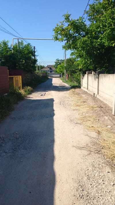 The image shows a dirt road in the village of Mitoc, Moldova. The road is lined with trees and fences, and there is a house in the distance. The sky is clear and blue. The image appears to have been taken on a sunny day.