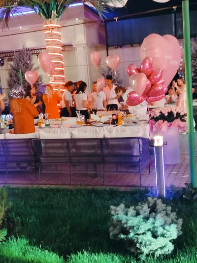 The image depicts an outdoor evening gathering or party, possibly a celebration, at a venue resembling a restaurant or event space. A long table is set with white tablecloths, place settings, and drinks, suggesting a dining or reception area. A cluster of pink heart-shaped balloons, including a large one with "MISS NAIMA" written on it, is prominently displayed, indicating a significant event, perhaps a birthday or a graduation.
Several people are visible, dressed in semi-formal attire. Some are standing around the table, possibly interacting with each other, while others appear to be in the background. A woman in an orange dress and a man in a white shirt are among those in the foreground. The setting features decorative elements like a palm tree wrapped in glowing lights and a large floral arrangement in a white container. The overall ambiance is festive, with artificial lighting contributing to the atmosphere. The scene appears to be located in Aksukent, Kazakhstan, based on the provided context.