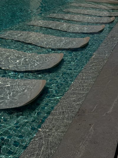 A high-angle shot shows a shallow swimming pool with stepping stones at El Gouna, Egypt. The pool's water is clear with a slight ripple. The pool tiles appear dark teal with visible grout lines. Gray stepping stones are semi-submerged in the pool, arranged in an arc. Adjacent to the pool is a gray concrete walkway, slightly wet from the water. Sunlight refracts through the water, creating ripple patterns on the stones and the pool floor.