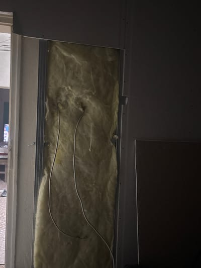 The image shows an unfinished interior wall, likely in a residential or commercial building under renovation. The wall cavity is filled with yellow fiberglass insulation, and white electrical wires are visible running through it. A metal stud frame is also present, outlining the cavity. To the left, a doorway leads to another area, where part of a television and a pair of shoes can be seen. To the right, a piece of drywall is leaning against the wall. The lighting is dim, suggesting the work is being done in low light conditions or at night. The scene does not provide any specific visual cues to pinpoint its location in Krushevo, Bulgaria.