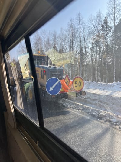 This image captures an outdoor scene in Pesterevo, Russia, viewed through the window of a vehicle. The perspective includes a window frame and part of a curtain on the left. The central subject is a yellow and gray 'SHANTUI' branded heavy construction vehicle, identifiable as a front-end loader or road clearing equipment, positioned on the right shoulder of an asphalt road. The vehicle displays an amber rotating beacon on its roof and illuminated red lights on its rear. Affixed to its rear are a blue circular mandatory sign with a white diagonal arrow pointing downward-left, a yellow triangular warning sign (partially obscured), and a yellow circular speed limit sign indicating "50". An orange high-visibility safety vest is draped over the equipment and signs.
The vehicle is actively engaged in pushing or clearing snow and slush from the road surface, creating distinct patterns of disturbed snow along the roadside. To the immediate right of the road, a substantial bank of accumulated snow is visible. The environment consists of a forest in the background, primarily composed of bare deciduous trees with some interspersed evergreen conifers, set against a clear blue sky. No individuals are explicitly visible within the frame.