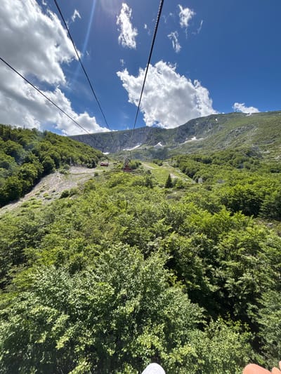 The image depicts a view from a ski lift ascending a lush green mountain landscape in Motički Gaj, Montenegro. The ski lift cars, painted red, are visible ascending the slope. The surrounding environment is characterized by dense green trees, rocky terrain, and a towering mountain in the background. The sky is a vibrant blue, dotted with white, fluffy clouds, and the sun casts a bright glare, creating lens flare effects. The image captures a sense of natural beauty and outdoor recreation in a mountainous setting.
