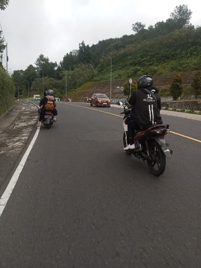 The image displays a multi-lane asphalt road scene under an overcast sky, located in Saing Ta Mawt Lay, Myanmar. The road features a solid white line marking the left shoulder and a dashed yellow line delineating lanes. Two motorcycles are present in the midground, traveling away from the viewer. The motorcycle on the left carries two individuals; the rear passenger wears a light brown backpack and dark clothing. The motorcycle on the right, positioned closer to the viewer and more centrally in the right lane, is operated by a single person wearing a black helmet, a black jacket with "3/4 DESIGN" written in white on the back, black pants, and white shoes. Further ahead on the road, an orange-red compact car is visible in the right lane, and a white van appears in the distant left lane. The left side of the road is bordered by a concrete curb and a wall covered in green ivy. To the right, a steep hillside densely covered with green trees and undergrowth rises, featuring visible concrete retaining wall sections. Streetlights are spaced along the road.