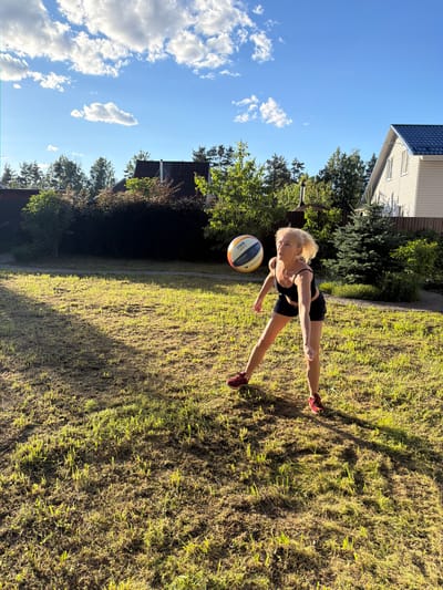 A blonde woman in athletic wear is playing volleyball in a grassy yard in Vaskelovo, Russia. The woman is in mid-action, with her right arm raised, about to hit a volleyball. The ball is in the air above her head, with the FIVB logo visible. She is wearing a black sports bra and shorts, and red athletic shoes. The yard is well-kept, with green grass and a few shadows cast by trees. In the background, there are houses, trees, and a fence. The sky is blue with a few white clouds, indicating it is a sunny day. The overall scene is one of outdoor recreation and leisure.