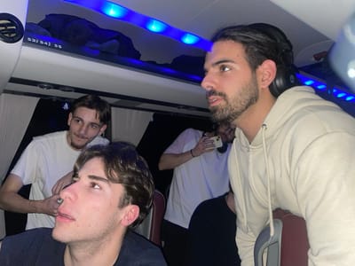 Four young men occupy the frame within a bus-like setting. One man wears a beige hoodie and large headphones. Another man in the foreground wears a dark shirt. A third, with a mischievous look, wears a white shirt and is partly obscured. The fourth man is in the background, also wearing a white shirt and looking at his phone. Blue lights illuminate the ceiling above, and some bus fixtures are visible.