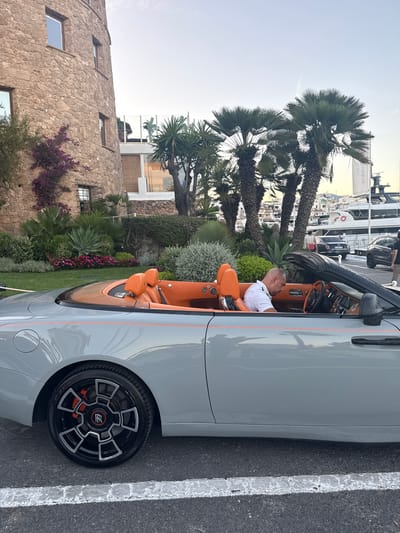 The image shows a man sitting in the driver's seat of a gray convertible car with an orange interior. The car is parked outdoors next to a paved area with a white line. Behind the car, palm trees and lush greenery are visible. In the background, there is a stone building with windows and a modern building with large glass windows. Further in the distance, several yachts are docked at a marina under a clear, pale sky, suggesting a daytime setting, likely late afternoon given the soft light. The man appears relaxed, looking forward. The scene suggests a luxurious leisure environment, possibly a coastal resort. No visible text is clearly legible.
