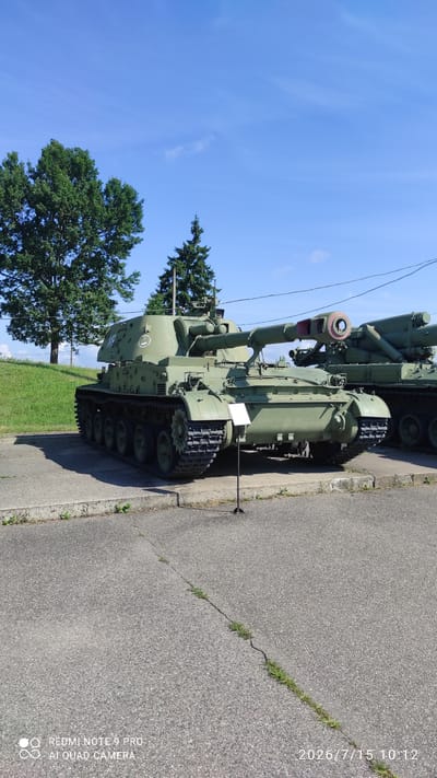 An outdoor shot shows two olive green self-propelled artillery pieces parked on a concrete platform next to an asphalt road. The foreground vehicle is a tank with a long barrel and treads. A white sign is placed in front of it. The background vehicle is partially obscured. Lush green grass and trees are visible behind the concrete platform, under a clear blue sky with a few wispy clouds. Power lines are visible in the upper right background. The timestamp on the image reads "2026/7/15 10:12". The lower-left corner displays "REDMI NOTE 9 PRO AI QUAD CAMERA". The location is identified as Лашанскі сельскі Савет, Belarus.