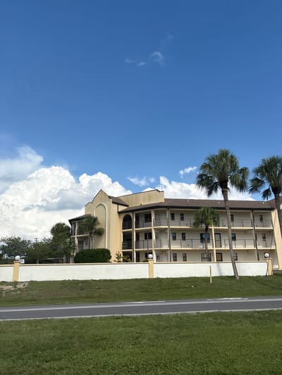 In North Port, Florida, USA, a three-story building with a pale yellow facade stands adjacent to a white stucco wall. The building features multiple balconies with black railings on each level. Several palm trees are visible in front of the building, with one tall, slender palm tree positioned prominently in the foreground. The sky above is a clear blue, punctuated by large, fluffy white clouds. In the foreground, a paved road with double white lines runs through a grassy area.