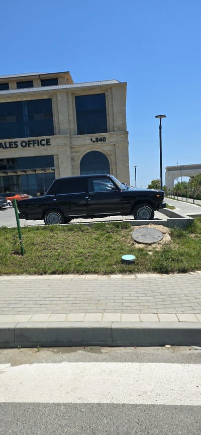 The image shows a black sedan parked on a street next to a modern building. The building has a light-colored facade with dark tinted windows. The words "SALES OFFICE" are visible on the building, along with the number "840". A street lamp is also visible in the background. The car is positioned parallel to the sidewalk, with its front facing towards the left side of the frame. The street is paved with light-colored bricks, and there is a strip of grass between the sidewalk and the road. The sky is clear and blue, suggesting a sunny day. The scene appears to be set in an urban or suburban environment. There are no people visible in the image. The image is taken from a slightly elevated perspective, looking down at the car and the surrounding area. The orientation of the image is vertical.