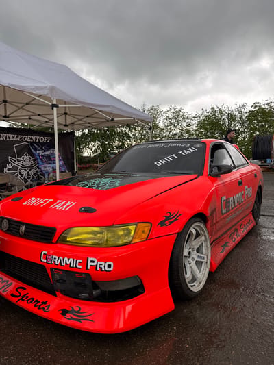A bright orange drift car is angled in the image, with a white tent partially visible to the left. The car features decals like "DRIFT TAXI" and "CERAMIC PRO," along with tribal flame designs. It's parked on wet pavement, suggesting a recent rain. The sky is overcast with grey clouds. A person is visible inside the car. A banner that reads "NTELEGENTOFF" is partially shown. A tire is near the back of the car on the road. The photo was taken in Pavlovskaya, Russia.
