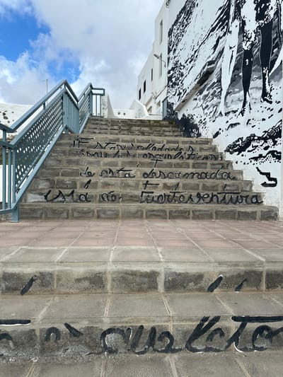 The image displays an outdoor concrete staircase ascending a hillside in Puerto del Rosario, Spain, under a partially cloudy sky. The staircase, constructed from light grey and brown block pavers, features dark grey metal railings with perforated panels on its left side, extending upwards and out of frame. Black handwritten script is painted across the risers, partially legible as "Si estás cansado y orgulloso de esta descarada vida en Fuerteventura" (If you are tired and proud of this shameless life in Fuerteventura). To the right of the stairs, a multi-story white building facade is adorned with a large black and white mural depicting stylized human legs and an abstract, textured background. A street light is mounted on the white building wall above the mural. The ground level in the foreground consists of square reddish-brown and grey paving tiles. No individuals are present in the scene.