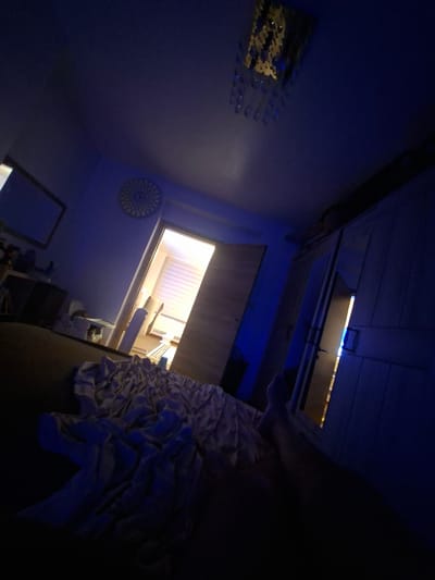 The image depicts an interior scene, possibly a bedroom, in Evere, Belgium. The room is dimly lit with blue ambient lighting and a bright light emanating from an open doorway in the background. A crumpled duvet or blanket is visible in the foreground, partially obscuring a person's legs and feet, suggesting someone is lying down.
The room's features include a ceiling light fixture with crystals, a circular decorative clock on the wall, a wardrobe with mirrored doors, and a framed mirror on the wall. The doorway in the background leads to a room with blinds on a window and what appears to be furniture. The overall impression is of a private, potentially domestic, space. The tilted angle of the photograph adds a dynamic, though disorienting, perspective to the scene.