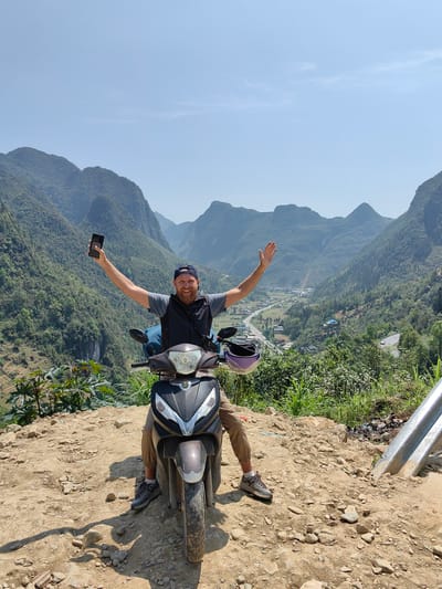 A medium, outdoor shot shows a man on a parked scooter, with a mountain vista in the background. The man is fair-skinned with a beard and a blue baseball cap. He wears a gray T-shirt, a black vest, and tan pants. He is sitting on a brown scooter, facing forward with his arms raised in a celebratory gesture. In his left hand, he holds a black smartphone. A purple helmet sits atop the scooter’s front.
The man is positioned on a rocky, dirt path. Behind him, the scene opens up to a vast valley with layered mountains covered in lush greenery. A small village can be seen in the valley below. The sky is clear and light blue.
The setting is Phố Bảng Commune, Vietnam.
