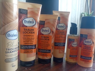 In the city of Aidemir, Bulgaria, on a brown, wooden surface, a collection of Balea hair care products is displayed. The products, all in orange and cream packaging, are arranged from left to right. The leftmost item is a bottle of "Blond Trocken Shampoo" with "keine weißen Rückstände nach dem Ausbürsten" (no white residue after brushing) and "lang anhaltende Frische" (long-lasting freshness) written on it, along with a vegan logo. Next to it are three tubes from the "Traum Locken" (Dream Curls) line. The first tube, labeled "SHAMPOO," also states "BIS ZU 24H DEFINIERTE LOCKEN*" (Up to 24h defined curls*) and "OHNE SILIKONE" (Silicone-free). The second tube is for "2IN1 OIL-IN-PFLEGE" (2in1 Oil-in-care), described as "Pflegt und definiert Locken mit intensiver Feuchtigkeit als Maske und Leave-In" (Cares for and defines curls with intensive moisture as a mask and leave-in). The third tube is a "SPÜLUNG" (Conditioner), also with the "BIS ZU 24H DEFINIERTE LOCKEN*" claim and "OHNE SILIKONE". A fourth item, a clear bottle with a blue pump dispenser, contains "TRAUM LOCKEN HAARÖL" (Dream Curls Hair Oil), noting "OHNE SILIKONE" and "PFLEGE-FORMEL MIT 5 WERT-VOLLEN PFLANZENÖLEN" (Care formula with 5 valuable plant oils). Finally, to the right of the hair oil is a tube of "TRAUM LOCKEN STYLING CREAM" with "Perfekt definierte, glänzende Locken und Hitzeschutz bis 220°C" (Perfectly defined, shiny curls and heat protection up to 220°C). All "Traum Locken" products in tubes and the styling cream claim to have a "PFLEGE-FORMEL MIT SHEABUTTER UND KERATIN" (Care formula with shea butter and keratin) and are suitable for "lockiges und gewelltes Haar" (curly and wavy hair). The background consists of a white curtain or blinds and a dark surface, suggesting an indoor setting.
