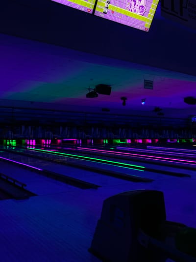 The image depicts a dimly lit bowling alley in Waynesboro, United States of America. The scene is illuminated by neon lights in shades of purple, green, and blue, creating a vibrant, almost psychedelic atmosphere. The bowling lanes are lined with these colored lights, and the pins at the far end of the lanes are also illuminated. In the foreground, a portion of a bowling ball return machine is visible, silhouetted against the colorful lanes. The ceiling is dark and features some hanging lights and projectors. A "CAUTION" sign is also visible on the ceiling. The overall impression is one of a modern, entertainment-focused bowling alley, likely operating at night or in low-light conditions. There are no people visible in the image.