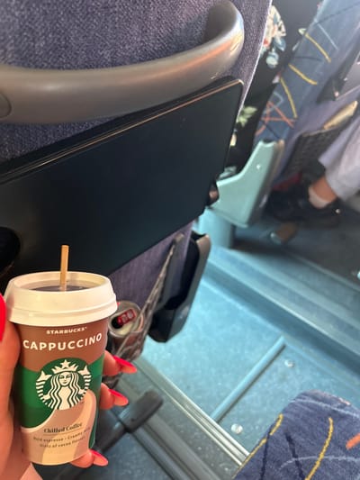 The image shows a person holding a Starbucks cappuccino in a disposable cup with a straw. The cup has the Starbucks logo and "CAPPUCCINO" written on it. In the background, there are seats that appear to be on a public transport vehicle. A can is visible next to the cup. The lighting suggests it is daytime. The location in Pirdop, Bulgaria cannot be determined from the image.