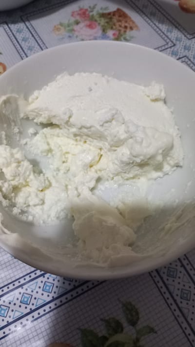 A close-up shot shows a white bowl filled with a creamy, white substance that appears to be ricotta cheese. The substance is textured, with some areas appearing smoother and others more clumpy. The bowl is placed on a table with a patterned tablecloth. The tablecloth has a floral pattern in the upper portion of the image and a geometric pattern in the lower portion. The scene is likely indoors. The location is not specified in the image or crops.