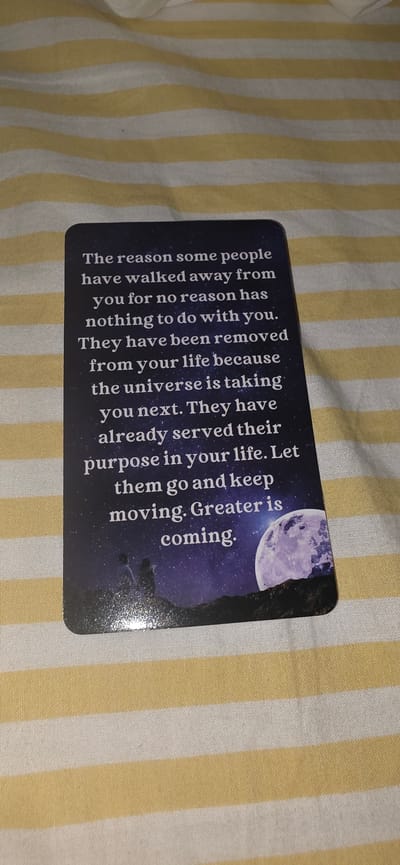 A close-up, angled shot shows a card lying on a striped fabric surface. The card is dark blue with white text and a night sky graphic. The text reads: "The reason some people have walked away from you for no reason has nothing to do with you. They have been removed from your life because the universe is taking you next. They have already served their purpose in your life. Let them go and keep moving. Greater is coming." The graphic on the right side of the card depicts a large purple moon against a starry night sky with silhouetted trees and figures in the foreground. The fabric beneath the card has alternating yellow and white stripes.