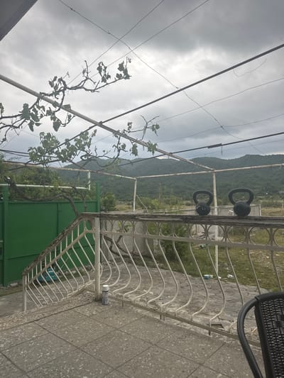 The image depicts an outdoor scene with a focus on a metal railing and a mountainous landscape under a cloudy sky. In the foreground, a light-colored tiled patio leads to a white metal railing with decorative, wavy vertical bars. A set of stairs with a similar railing is visible on the left, adjacent to a tall, green metal gate.

Two black kettlebells are placed side-by-side on top of the railing. Behind the railing, a wall or structure runs horizontally, and beyond that, lush green vegetation and trees are visible. In the background, rolling hills covered in dense forest rise towards the horizon, partially obscured by a heavy, overcast sky. The overall impression is one of a rural or semi-rural setting in Aragvispiri, Georgia, with the natural landscape being a prominent feature.