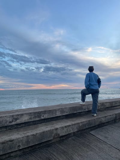 An individual stands facing west on a concrete promenade or seawall in La Grande-Motte, France, observing the sea. The subject wears a light blue long-sleeved sweater, dark trousers, and white athletic shoes; their right foot is elevated and placed on the upper concrete ledge of the multi-tiered structure. A dark fanny pack is worn diagonally across the subject's back. The sea exhibits a moderate chop, with a distinct splash visible where waves impact the concrete barrier on the left. The horizon is characterized by a heavily clouded sky displaying hues of blue, grey, pink, and orange, indicative of a sunset or late afternoon. A single white sailboat is visible in the distant sea to the right. The foreground consists of a textured concrete ground surface leading to two prominent horizontal concrete steps or barriers.