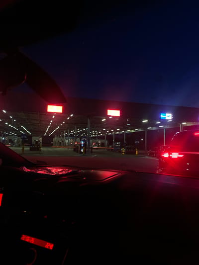 The image captures a view from inside a vehicle at a border crossing in Terespol, Poland, during nighttime. The sky is a deep navy blue, transitioning to a darker shade at the top. In the foreground, the dashboard of the car is visible, with a digital display showing the date "22/06/26" and the time "22:48". Red light from the dashboard illuminates parts of the interior.
The border crossing itself is a long, covered structure with numerous lanes marked by yellow poles and red lights, indicating a checkpoint. Overhead, bright fluorescent lights line the structure, illuminating the area. Red rectangular signs are mounted high up on the structure, and one larger sign displays blue and white graphics. Various booths and kiosks are visible along the lanes, presumably for border control. Several cars are lined up in the different lanes, waiting to pass through the checkpoint. The taillights of a dark-colored vehicle in the right lane are prominently illuminated with a bright red glow. The scene suggests a quiet, late-night atmosphere at an international border.