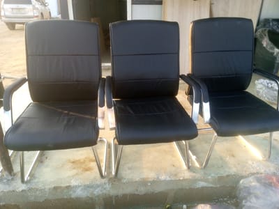 Three black executive chairs with chrome legs are arranged in a row on a concrete surface. The chairs have cushioned seats and backs with horizontal stitching. The background shows a silver SUV, a building with white doors, and some plastic-wrapped items. The scene appears to be an outdoor area, possibly a yard or a service bay, in Indo, Nigeria. The lighting suggests it is daytime.