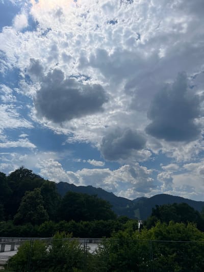 The image depicts a scenic view of the Oberammergau ski jump in Bavaria, Germany, set against a backdrop of lush green trees and a dramatic cloudy sky. The ski jump structure is visible in the distance, appearing as a slender white line ascending a mountainside. The surrounding mountains are covered in dense forest, with a clear ski slope visible cutting through the trees. The sky is filled with a dynamic mix of white and grey clouds, with patches of blue sky interspersed. In the foreground, there is a fence and some foliage, suggesting a viewing area or park. The overall scene captures a tranquil, natural landscape with a prominent man-made structure.
