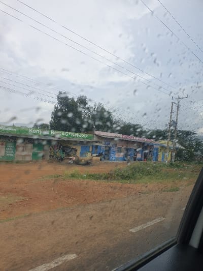 A rainy day view from a car window shows a roadside scene. Utility poles and wires stretch across the sky. A weathered, blue-painted building stands beside a dirt road. A green sign with "Kenya Best 4G Network" is visible. White lane markings are on the road, which is partially obscured by the rainy window.